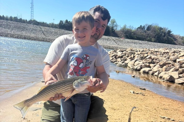 lake-texoma-fishing-report-march-2026-a Alt Text: Child holding striped bass with adult on shoreline near Denison Dam Lake Texoma during guided fishing trip